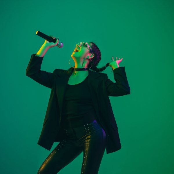 Caucasian female singer portrait isolated on green studio background in neon light. Beautiful female model in black wear with microphone. Concept of human emotions, facial expression, ad, music, art.